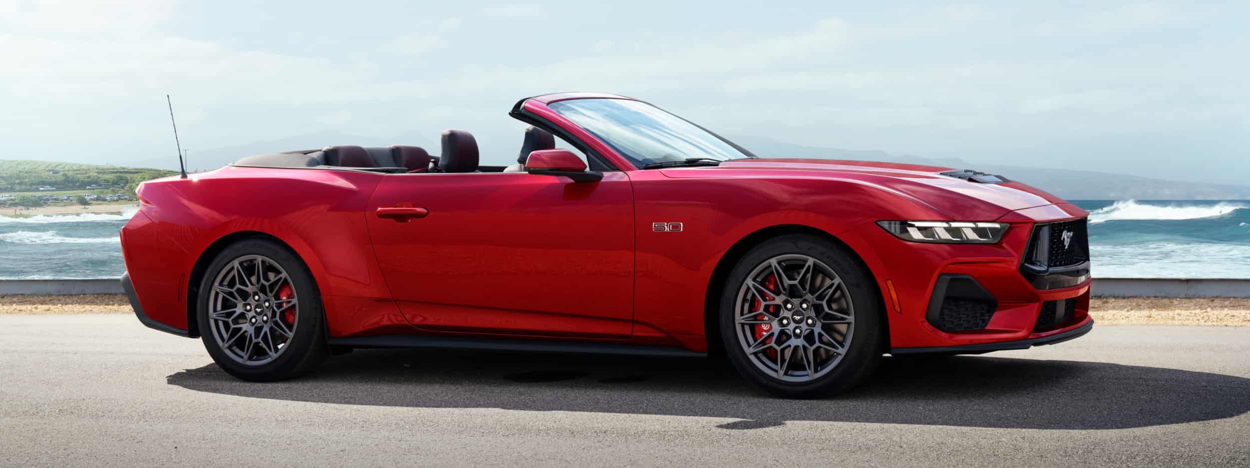 Red 2023 Ford Mustang 5.0 Convertible on a road by the ocean.