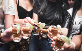 Five women standing in a circle holding shot glasses with alcohol and lime