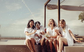 Four women sitting on a deck near a beach and opening a bottle of champagne