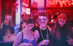 Three women share a drink in a bar while wearing decorative face masks