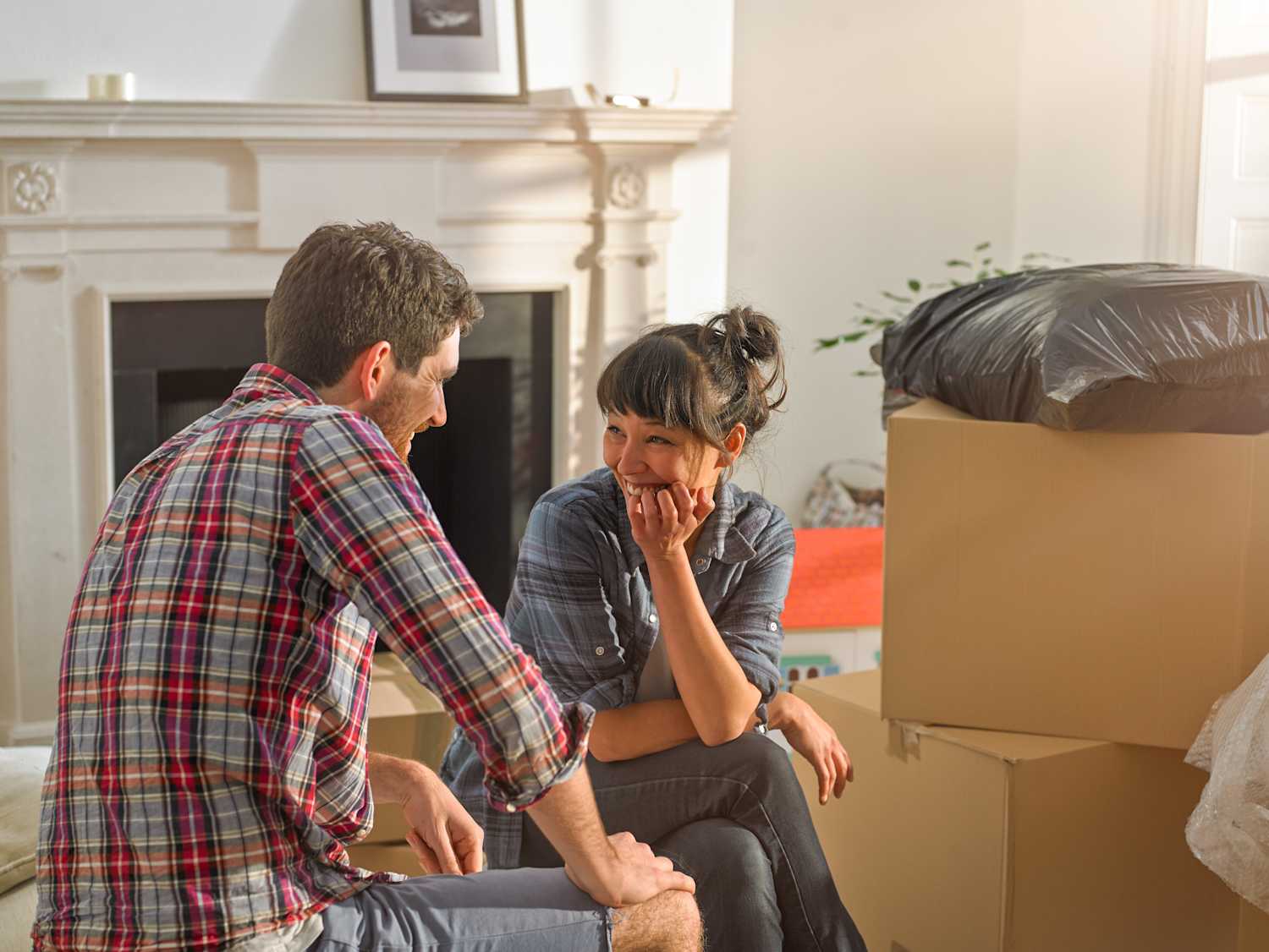 A happy couple sitting in a white lounge with a white fireplace surrounded by cardboard boxes