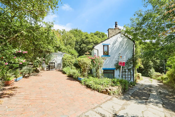 The exterior of a cute cottage in Pudsey with white walls and a driveway set into the hill, surrounded by trees and greenery