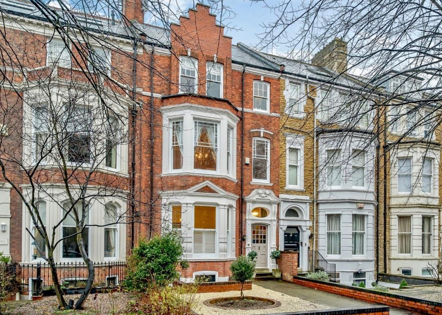 Large red brick Victorian townhouse with bay windows and a front garden with small shrubs and paved area