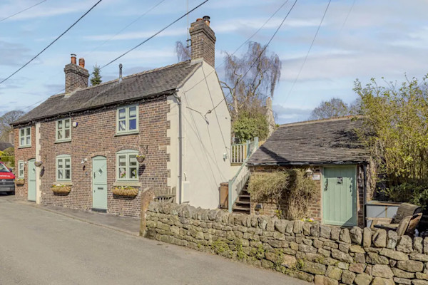 The exterior of a farmhouse-style cottage set close to the road with a low bricked wall in front of a small outhouse