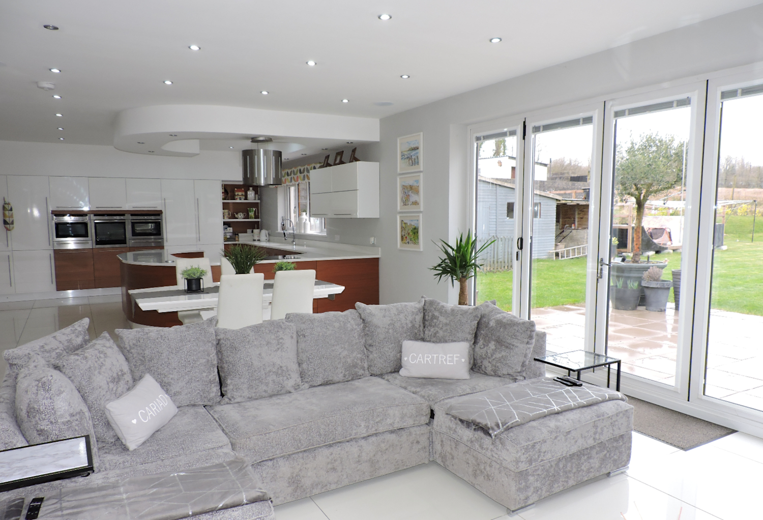 Kitchen and living area with modern units and a corner sofa, a white interior, and doors opening out into the garden