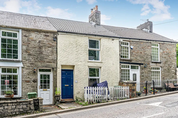 The exterior of a cream-coloured terraced cottage with a blue front door and two windows set close to the road in Wales