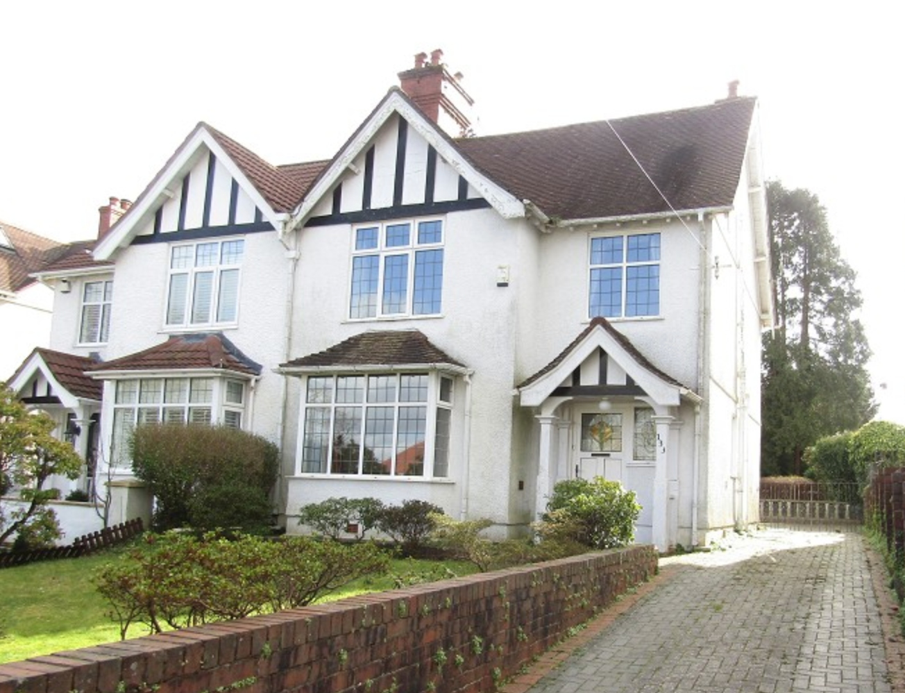 White house with pitched roof and mock Tudor beams, a porch area, drive and walled front garden laid to lawn