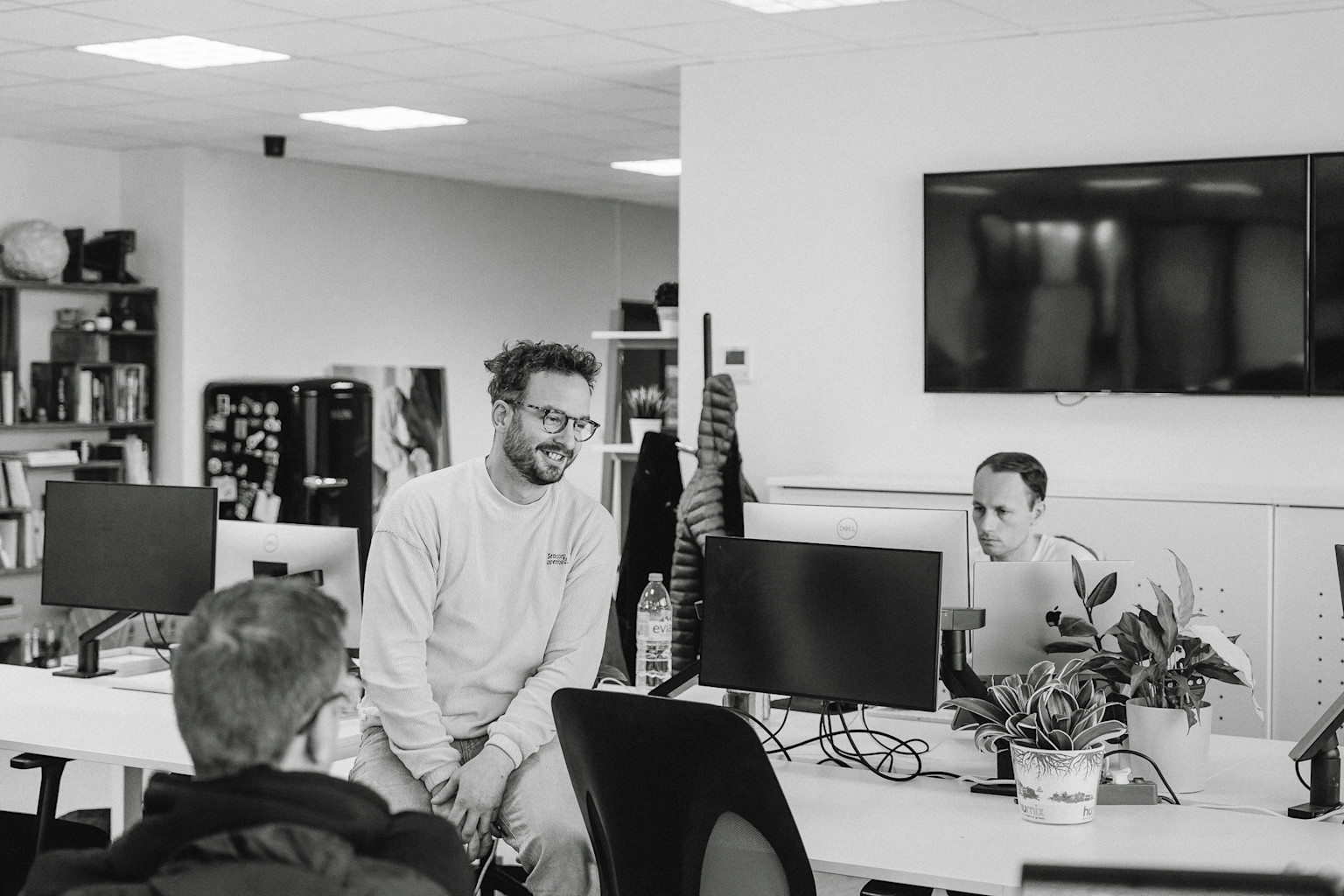 A picture of our colleagues having a conversation at the office with computer desks, potted plants, and a wall-mounted TV in black and white.