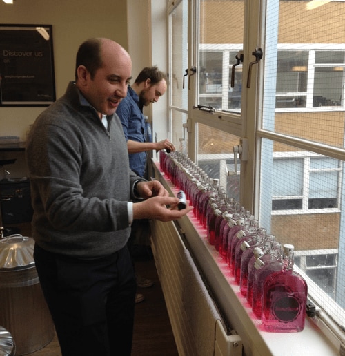 Hugo and Chris stand in front of a row of 30 to 40 bottles of gin on a window sill, adding pink food colouring.
