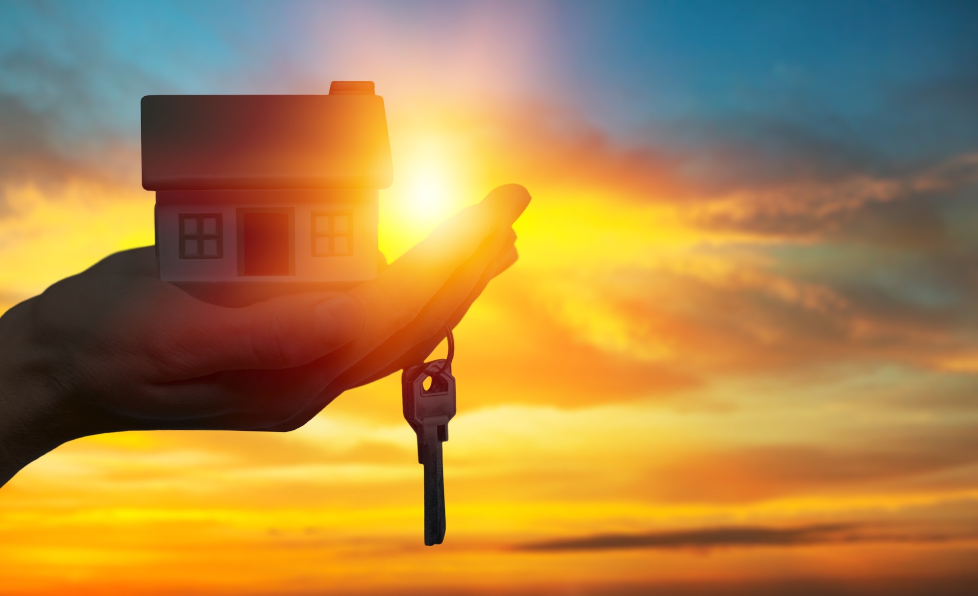 Miniature house model sits in the palm of a hand with keys dangling in front of a sunrise backdrop.