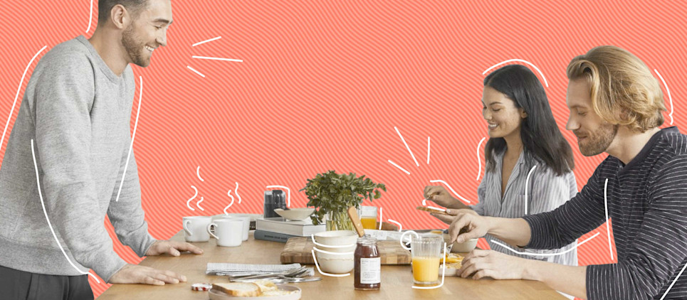 group up roommates, two males, one female gathered around a table for breakfast with orange. juice, coffee cups on a pink background