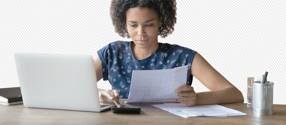 A woman sits at a desk with a laptop looking over paperwork.