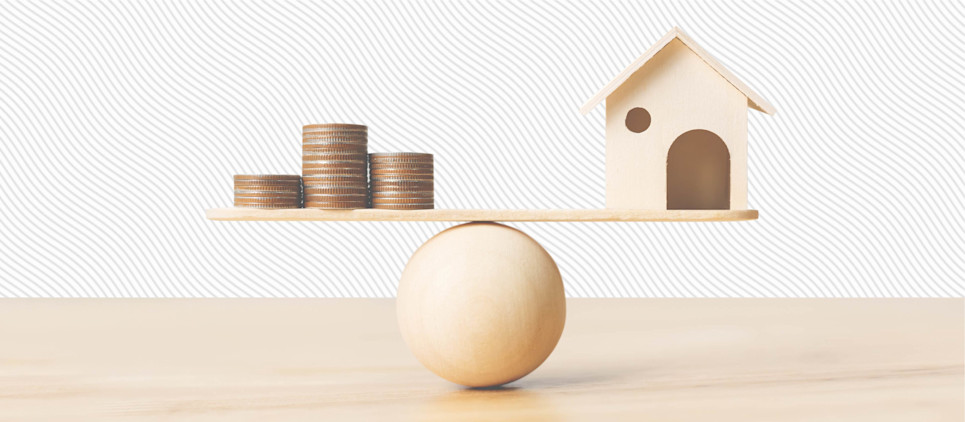 A wooden house figurine and stacked coins balance on different ends of a wooden seesaw.