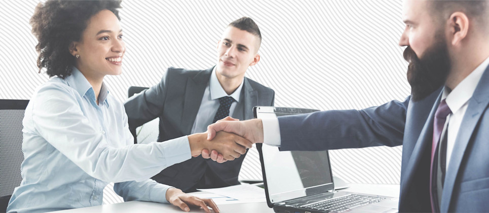 A woman shakes hands with a man sitting across from her at a desk while another man looks on.