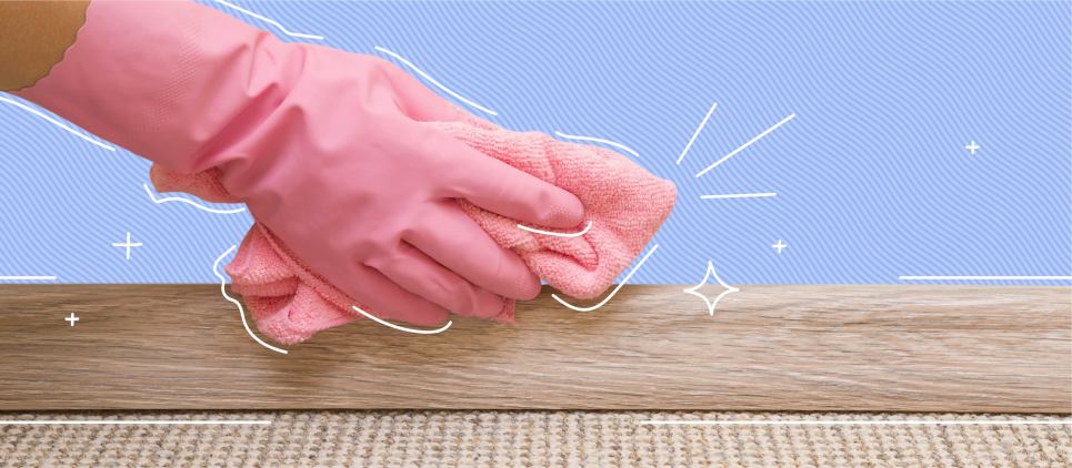 Hand with a pink cleaning glove scrubs a baseboard with a rag.
