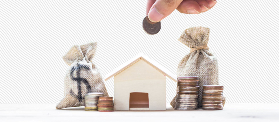 A hand holds a coin above a small model wooden house surrounded by sacks of coins.