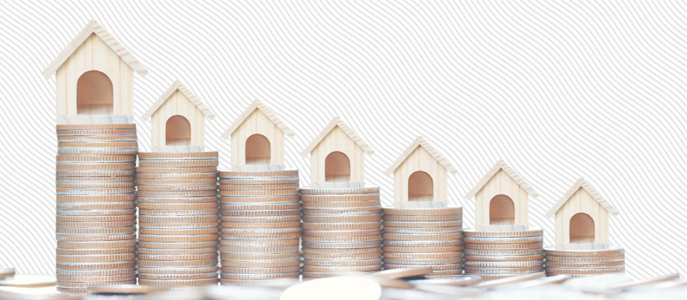 A row of miniature wooden house figurines sit atop stacks of coins. 