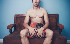 Man sitting naked on a sofa with a cushion covering himself