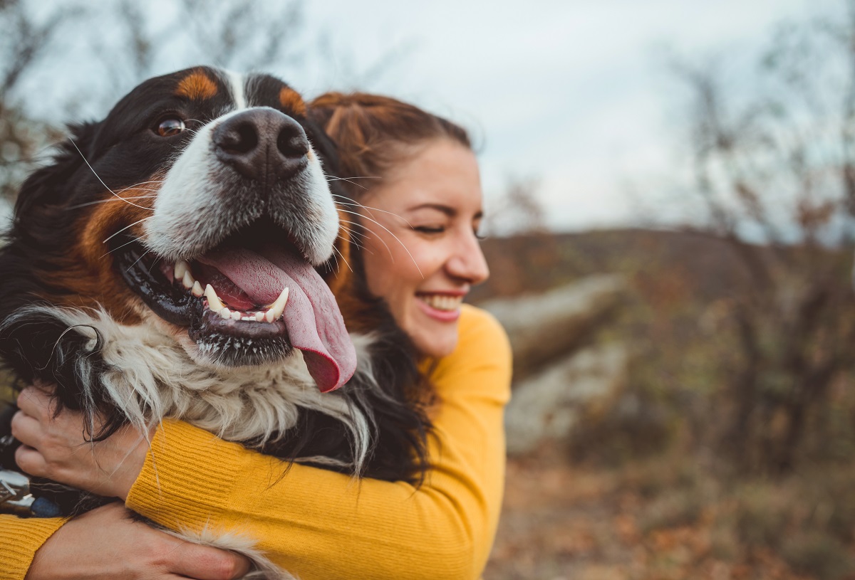 Picture of a woman with long brown hair and a yellow sweater embracing her large dog. She is excited about learning about benefits to improve her quality of life.