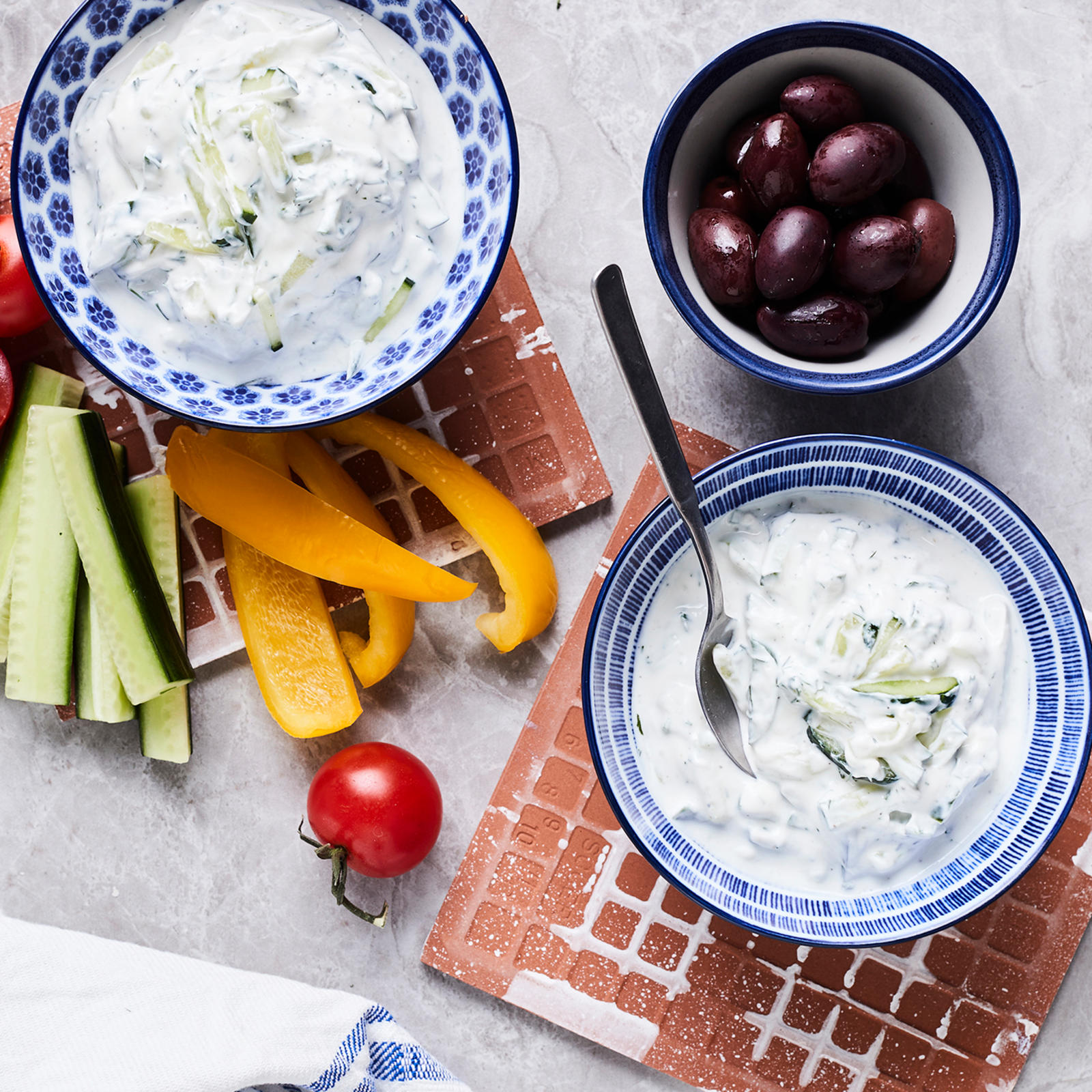 Tsatsikia kahdessa kulhossa, joista toisessa lusikka. Kulhojen vieressä pöydällä pilkottua paprikaa, kurkkua ja kirsikkatomaatteja sekä mustia oliiveja omassa kulhossaan.