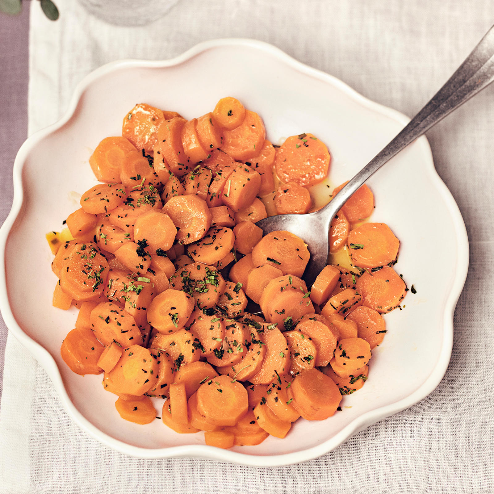 Rakuunaporkkanat vaalealla lautasella, jossa lusikka. Lautasen alla vaalea liina ja vieressä laseja, lautanen ja maljakko.