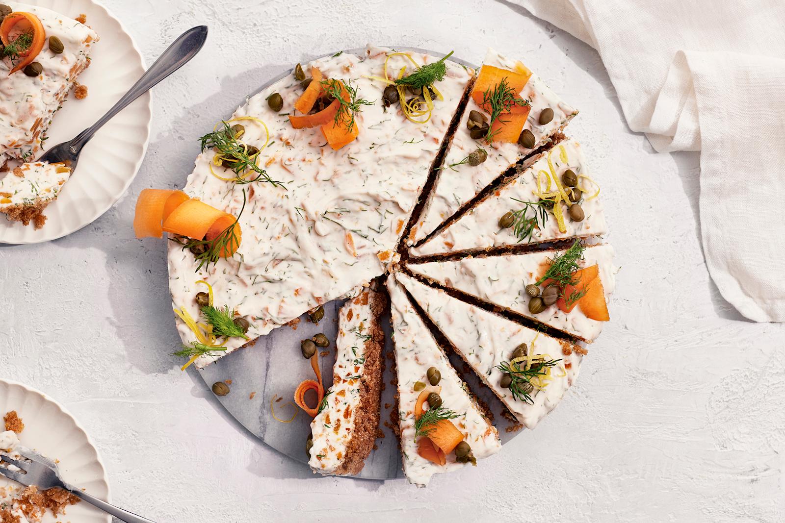 Vegaaninen kylmäsavulohijuustokakku, josta on leikattu useita paloja valmiiksi tarjoilulautaselle. Vieressä kaksi lautasta, joilla kakkupalat ja leivoshaarukat.