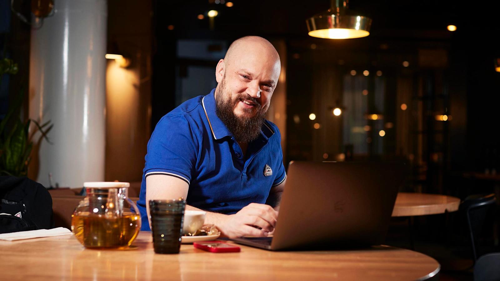 Artisti Pyhimys istuu pöydän ääressä. Pöydällä on kannettava tietokone, teekannu ja juomalasi.