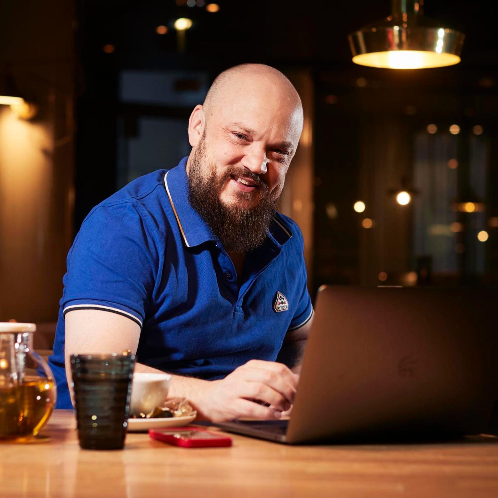 Artisti Pyhimys istuu pöydän ääressä. Pöydällä on kannettava tietokone, teekannu ja juomalasi.