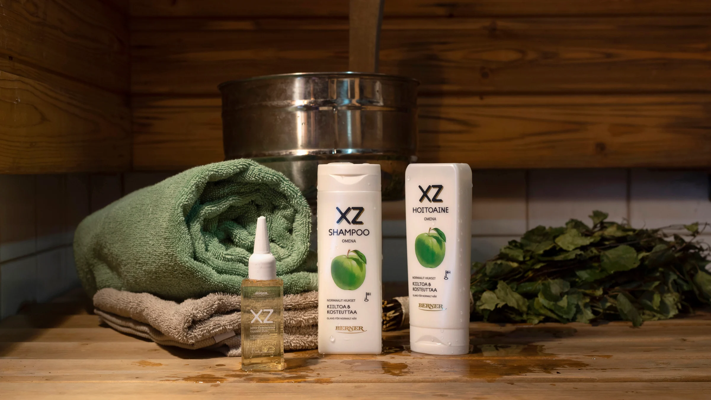 XZ-shampoo ja XZ-hoitoaine sekä hiusöljy kuvattuna saunassa. Taustalla pyyhe ja vihta.
