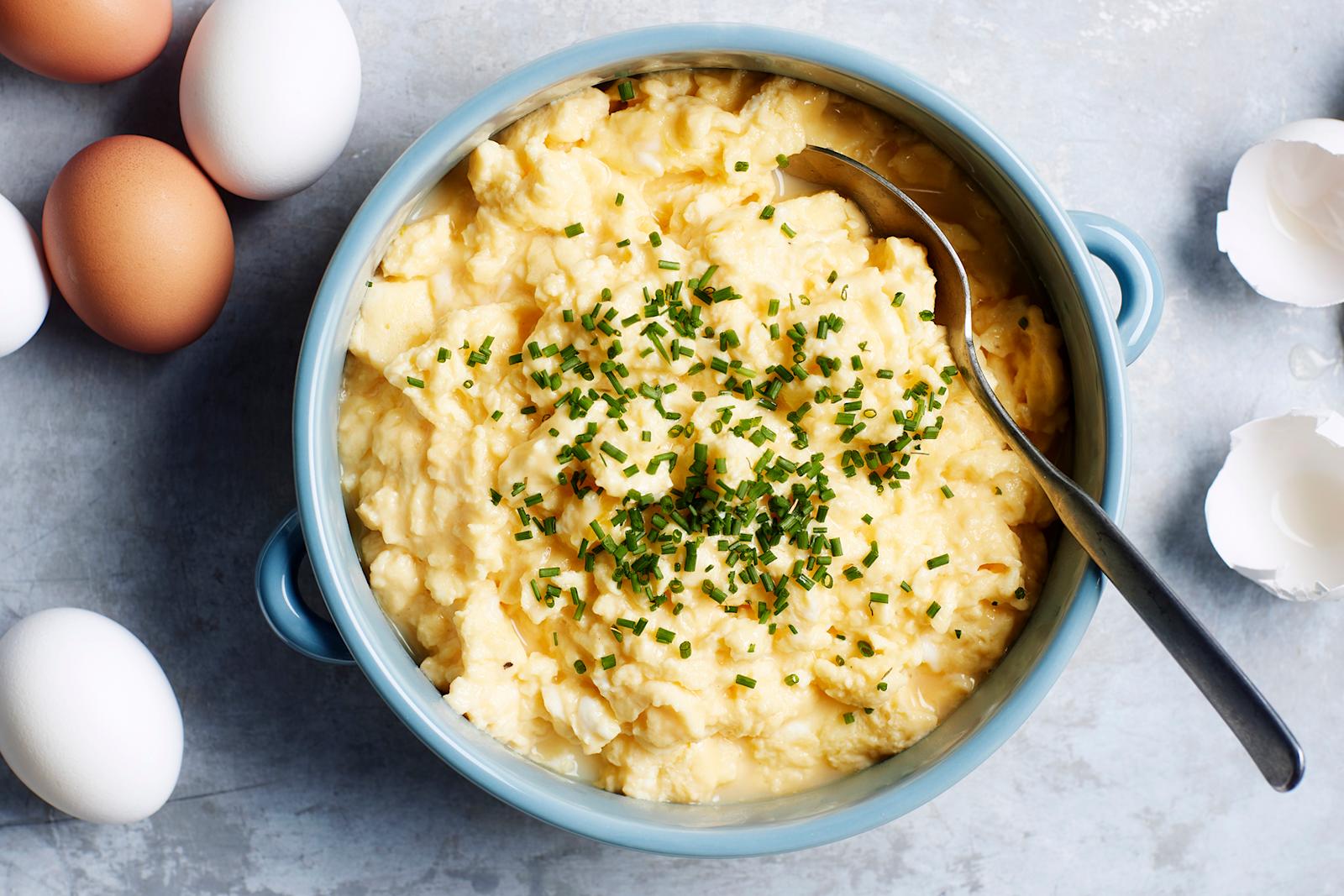 Munakokkelia ruohosipulilla koristeltuna vaaleansinisessä kattilassa. Kattilassa lusikka. Kattilan vierellä kananmunia ja kananmunan kuoria.