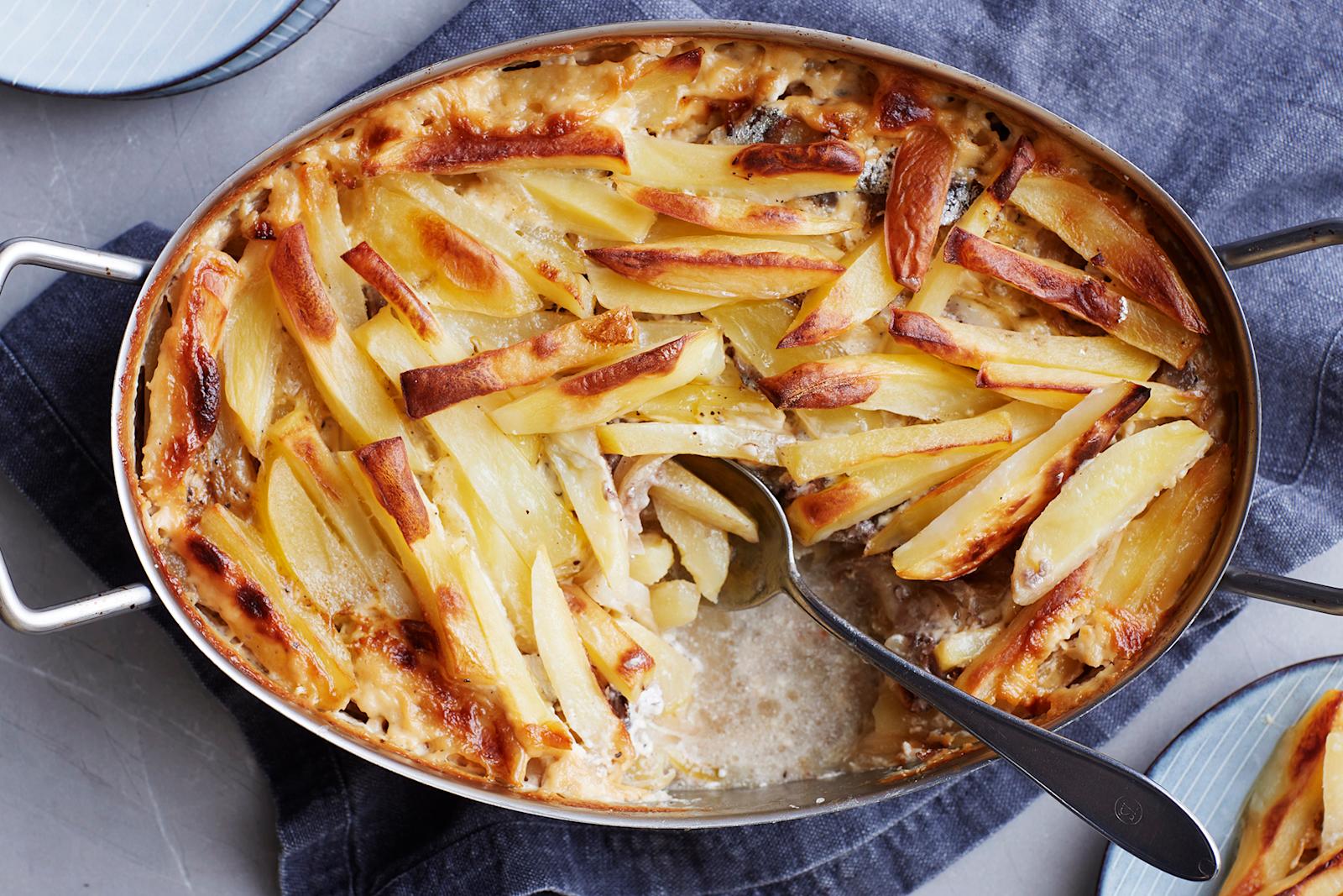 Janssoninkiusaus metallisessa uunivuoassa. Vuoassa tarjoilulusikka ja vuoan alla sininen liina. Kuvan laidoilla lautasia, joista yhdellä annos kiusausta.