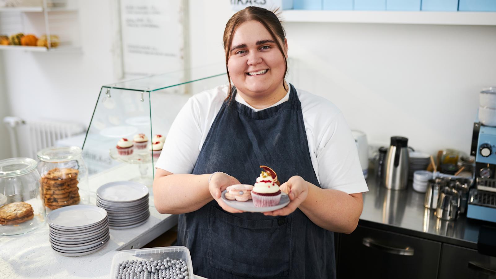 Erika Poussa pitelee käsissään leivoksia. Pöydällä on keksipurkki, lautasia ja karkkirakeita.