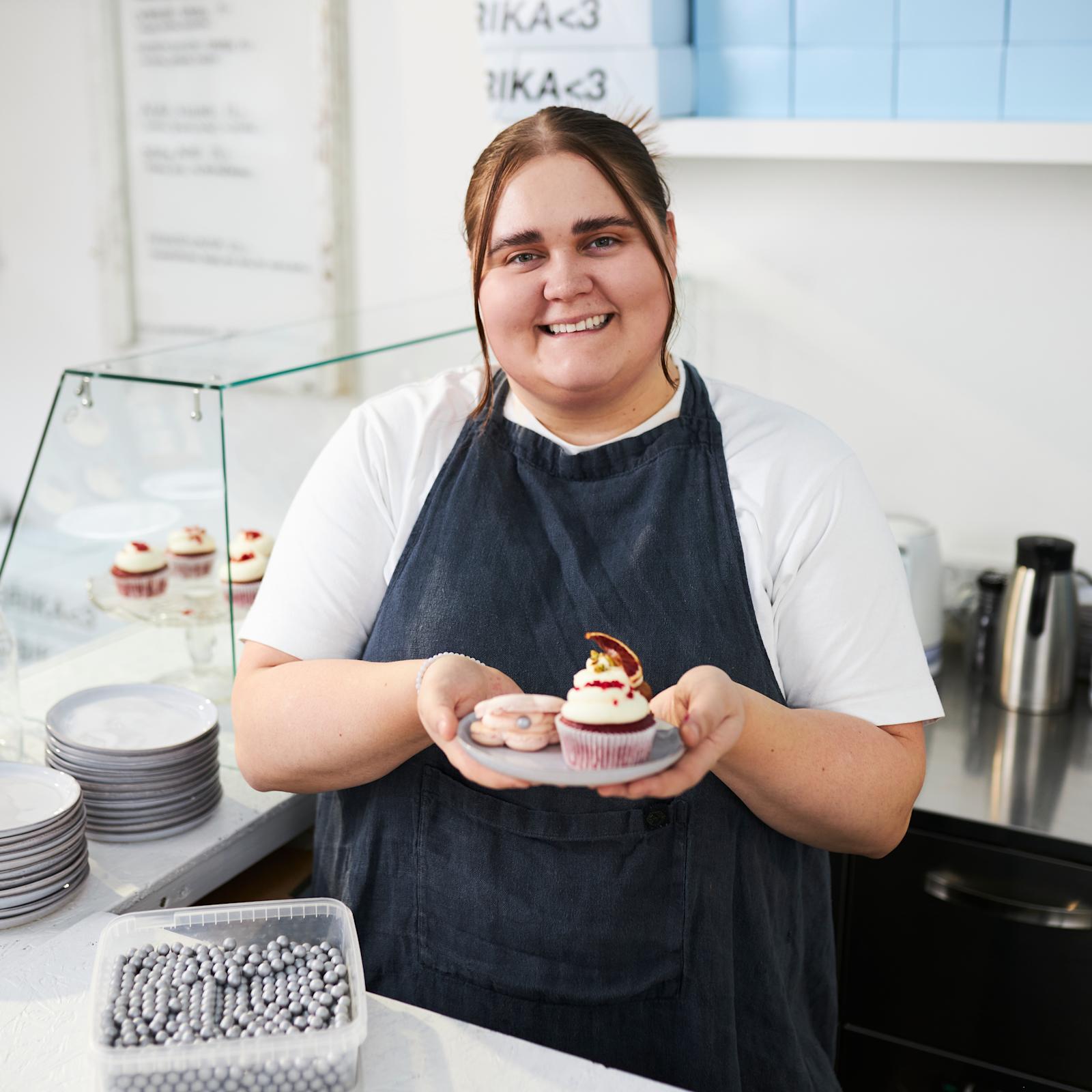 Erika Poussa pitelee käsissään leivoksia. Pöydällä on keksipurkki, lautasia ja karkkirakeita.