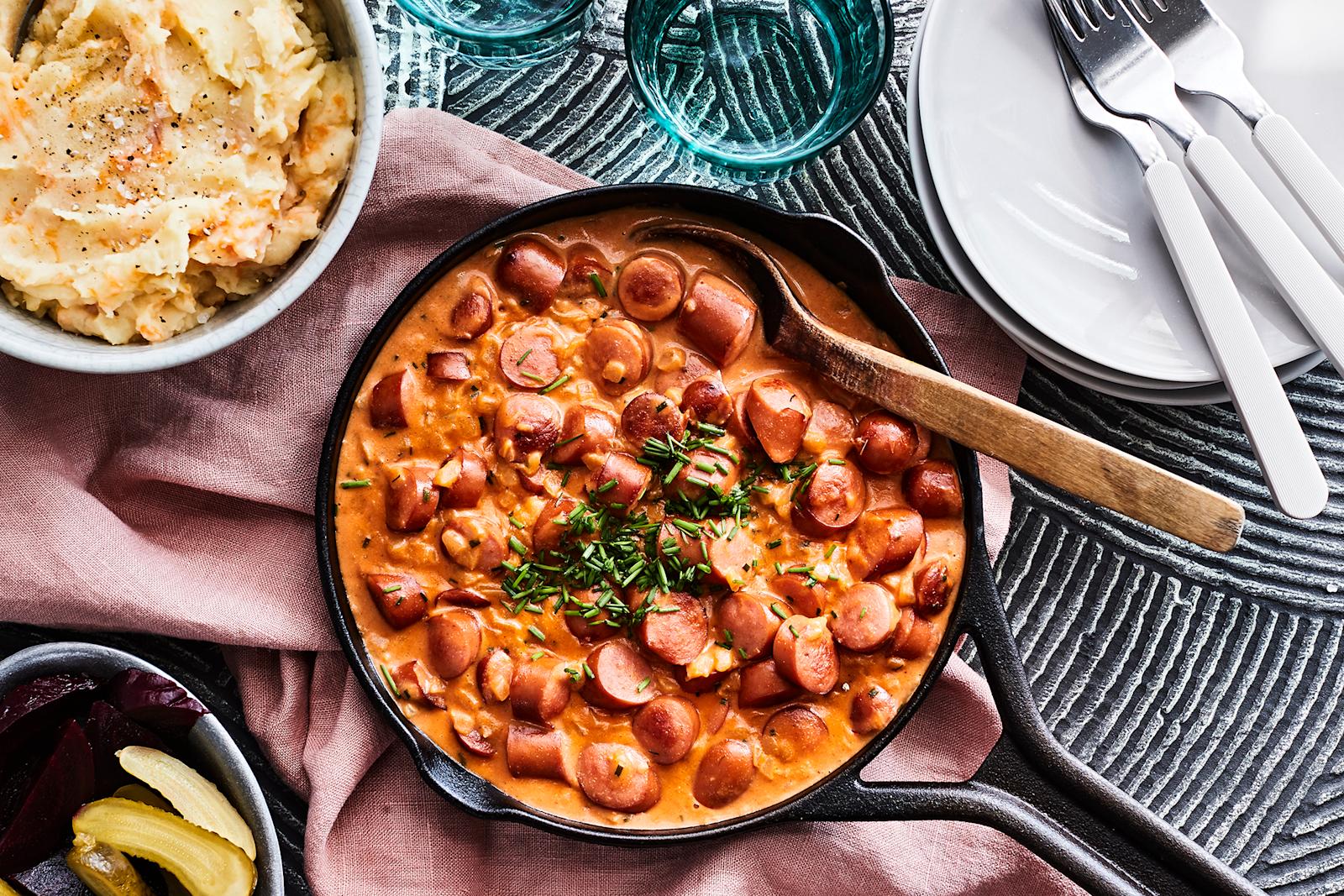 Nakkikastike paistinpannulla. Vieressä kulho juuresmuusia, maustekurkkuja ja punajuuria, pino valkoisia lautasia, kolme haarukkaa ja kaksi lasia.