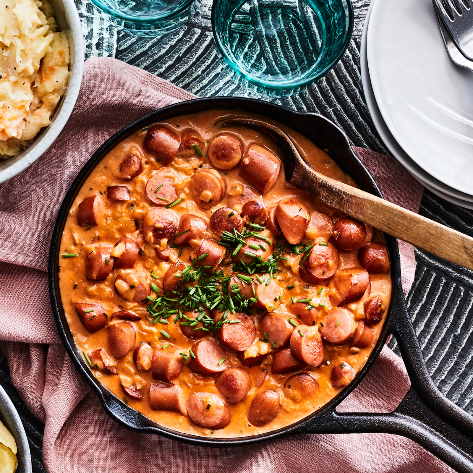 Nakkikastike paistinpannulla. Vieressä kulho juuresmuusia, maustekurkkuja ja punajuuria, pino valkoisia lautasia, kolme haarukkaa ja kaksi lasia.