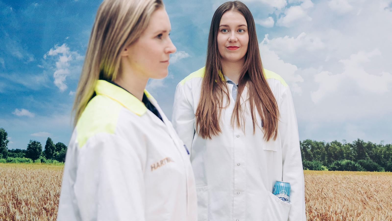 Kaksi naista seisoo peltokuvan edessä laboratoriotakeissa, toisen taskussa näkyy Long Drink -juomatölkki.