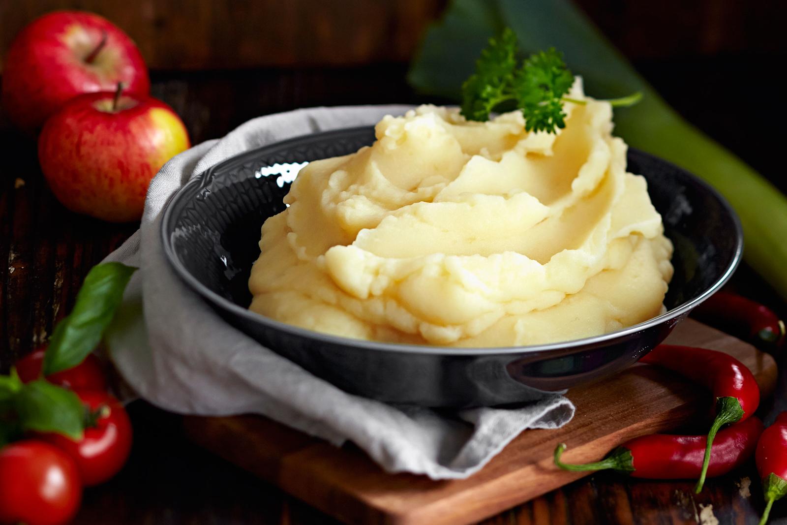 Perunamuusia tummassa tarjoilukulhossa, jonka alla liina ja puinen pannunalunen. Muusin päällä persiljatupsu. Kulhon ympärillä omenoita, tomaattia, chiliä ja purjosipuli.