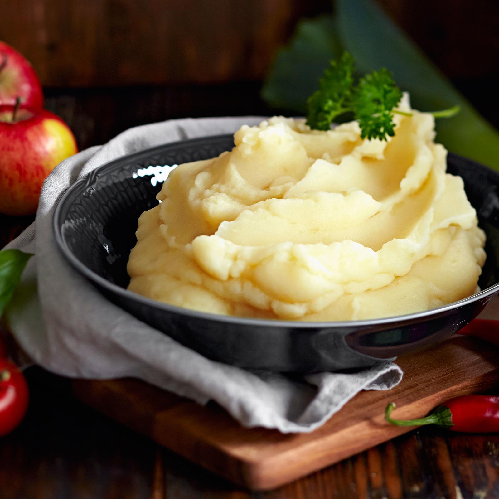 Perunamuusia tummassa tarjoilukulhossa, jonka alla liina ja puinen pannunalunen. Muusin päällä persiljatupsu. Kulhon ympärillä omenoita, tomaattia, chiliä ja purjosipuli.