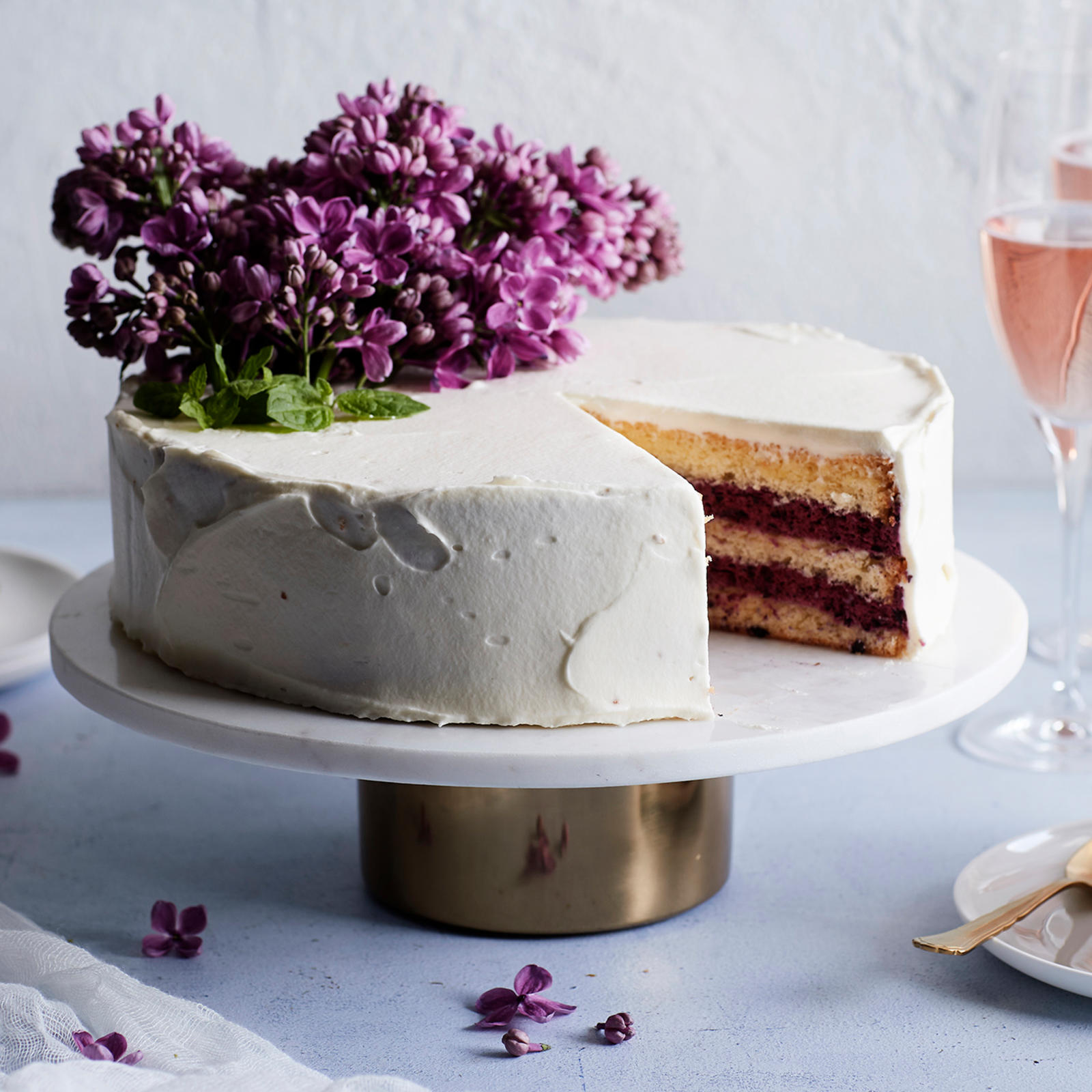 Täytekakku, joka koristeltu syreenin kukilla. Kakusta leikattu pala lautaselle. Kuvan taustalla kuohuviinilaseja ja lautanen, jonka päällä lusikoita.