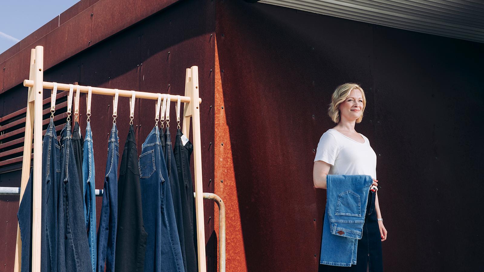 Farkkuja puisella rekillä, nainen kävelee pois päin rekistä käsivarrellaan taitellut farkut