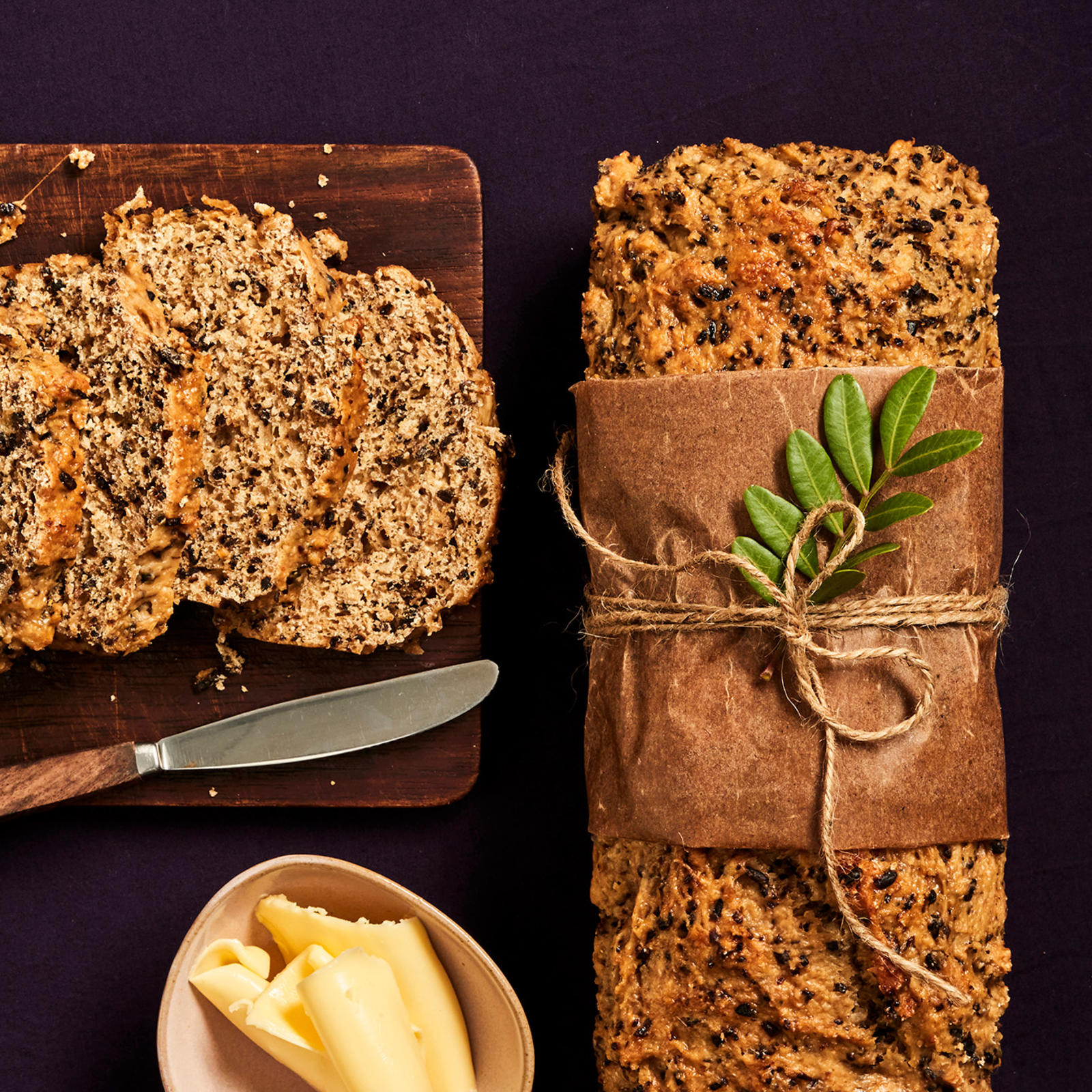 Saaristolaisleipä, jonka ympärille on sidottu pala leivinpaperia narulla. Naruun on kiinnitetty puolukan varpu. Vieressä viipaleiksi leikattu leipä leikkuulaudalla, leipäveitsi ja voikulho.