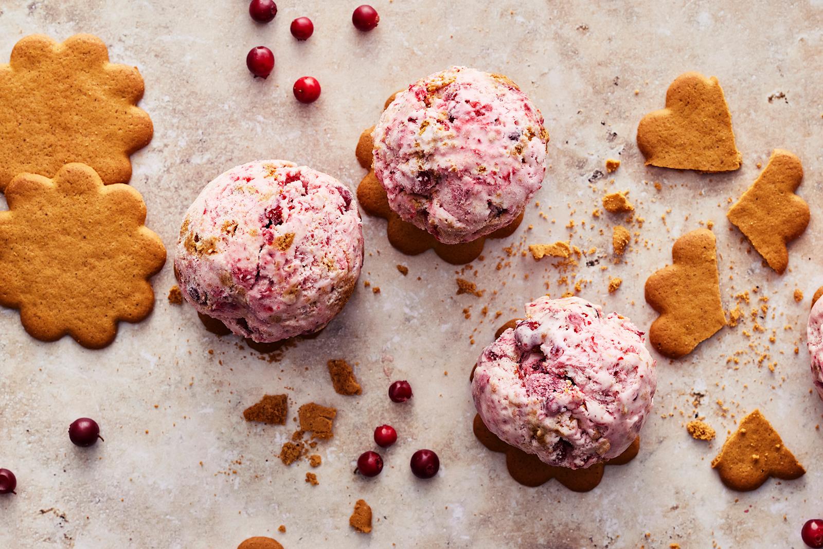 Neljä palloa piparkakku-puolukkajäätelöä piparkakkuje päällä. Pöytätasolla kokonaisia ja murennettuja piparkakkuja ja puolukoita.