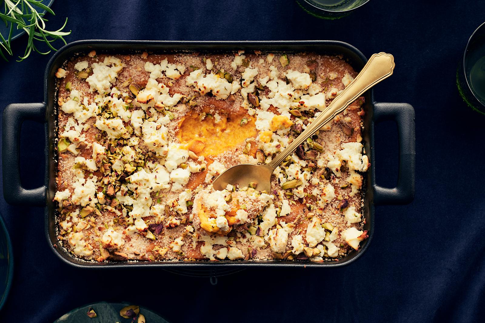 Feta-pistaasikuorrutteinen bataattilaatikko tummassa uunivuoassa. Vuoassa lusikka, jolla laatikkoa on kauhaistu.
