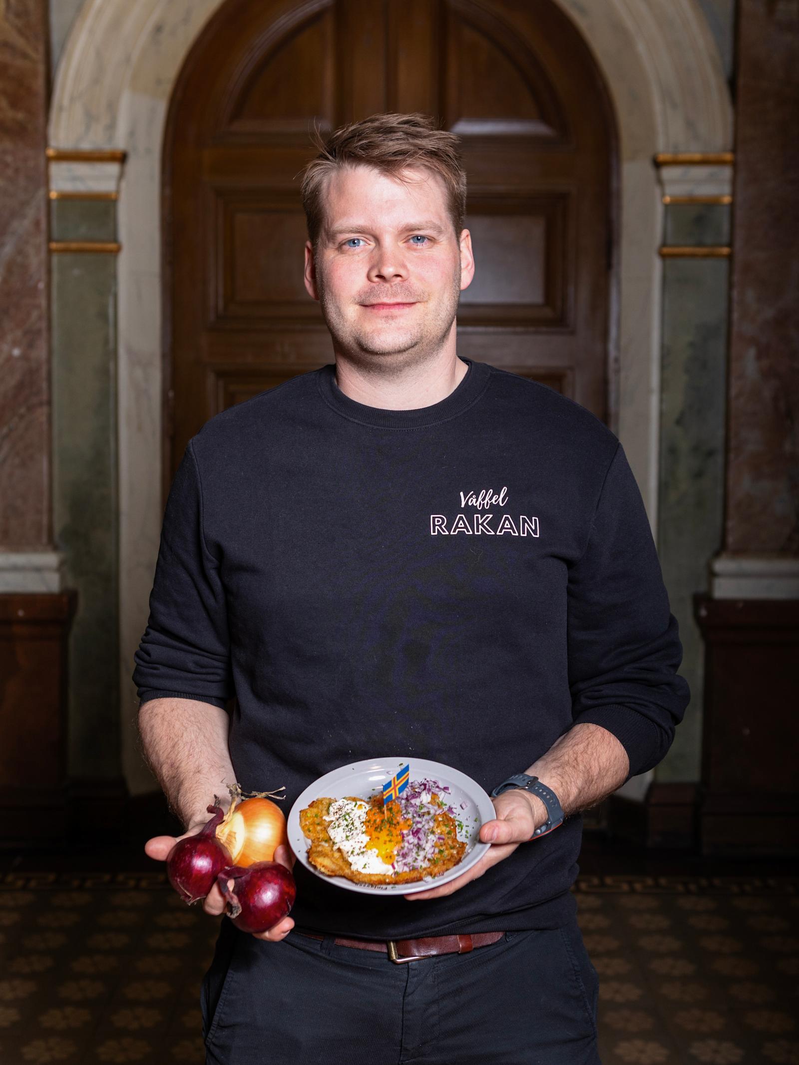 Jack Mattssonilla on käärinyt hihat mustasta paidastaan. Oikeassa kädessään hän pitelee kolmea sipulia. Vasemmassa kädessä puolestaan on lautanen, jolla on useammalla täytteellä kuorrutettu vohveli.