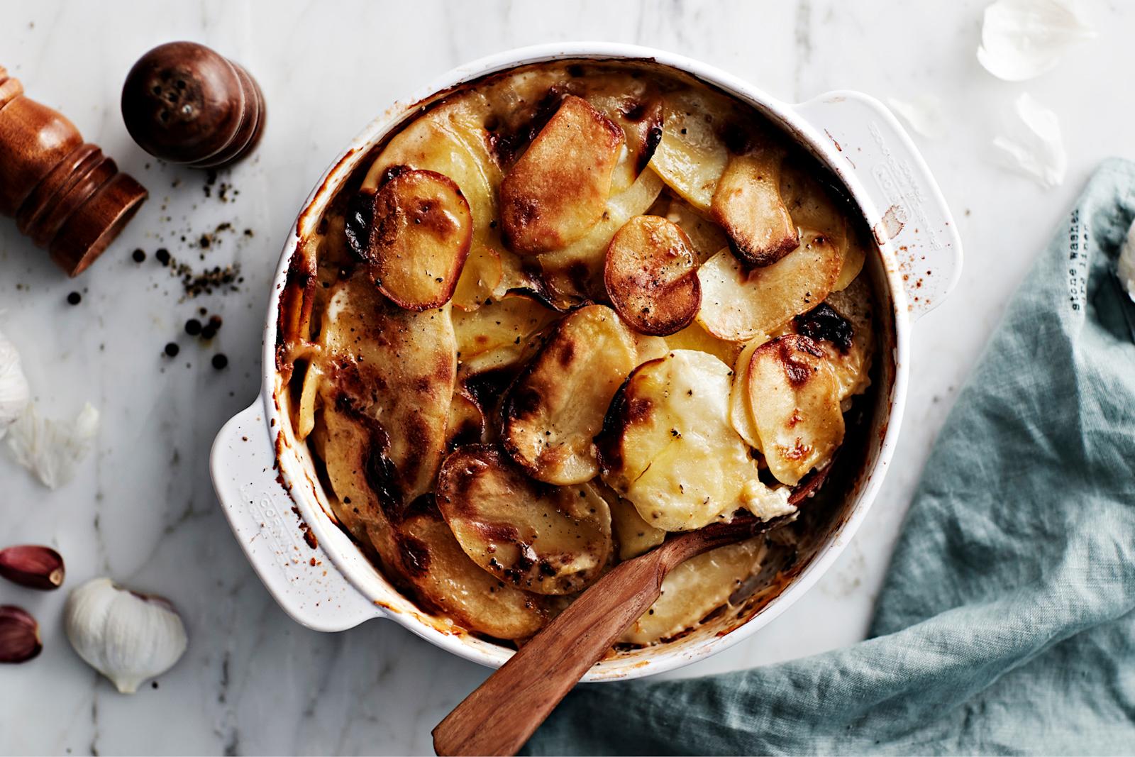 Kermaperunat valkoisessa uunivuoassa. Vuoassa puukauha, vieressä sininen liina, pippurimyllyjä ja valkosipuleja.