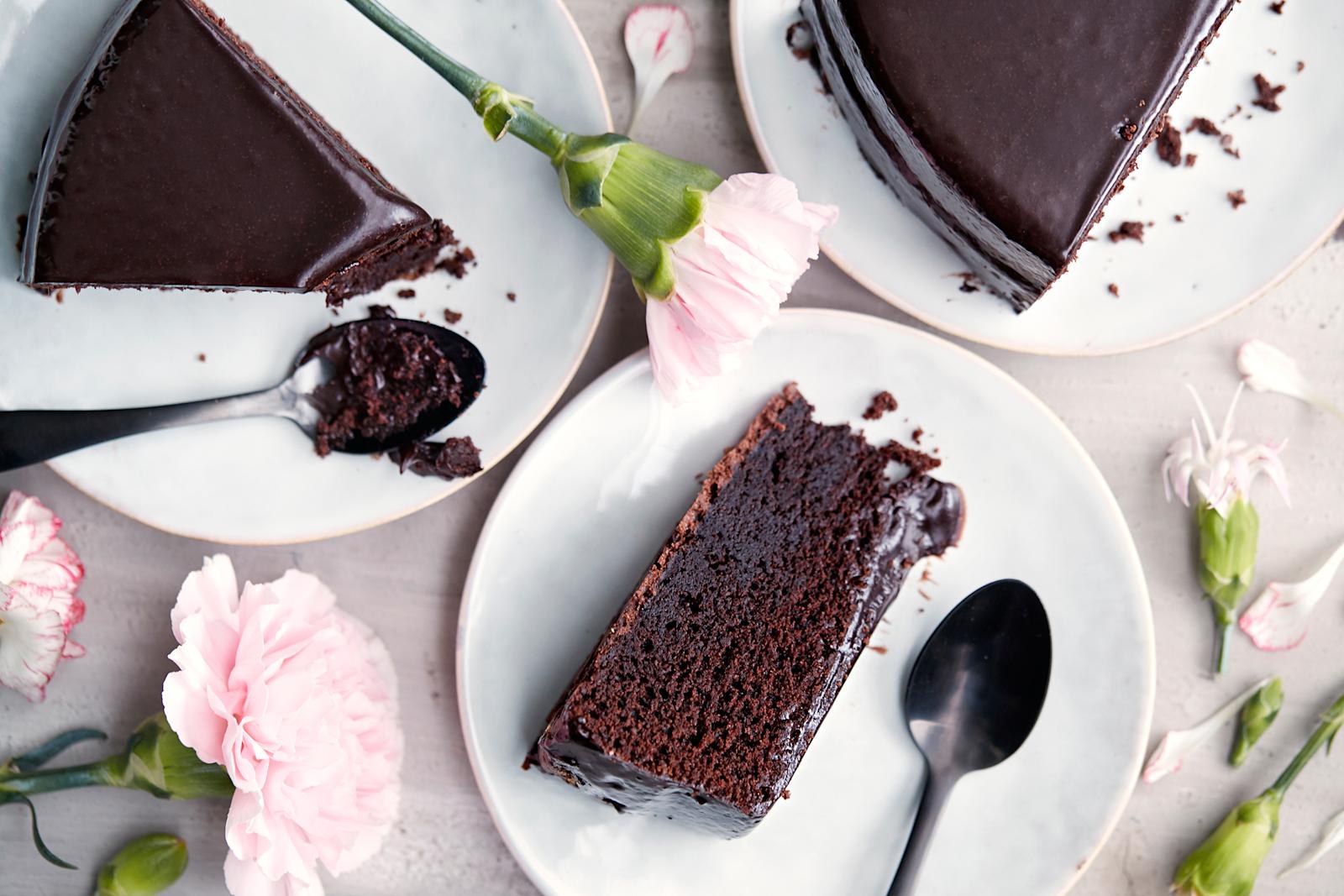 Kolme suklaakakkupalaa kakkulautasilla, kaksi lusikkaa ja vaaleanpunaisia neilikoita pöydän päällä.