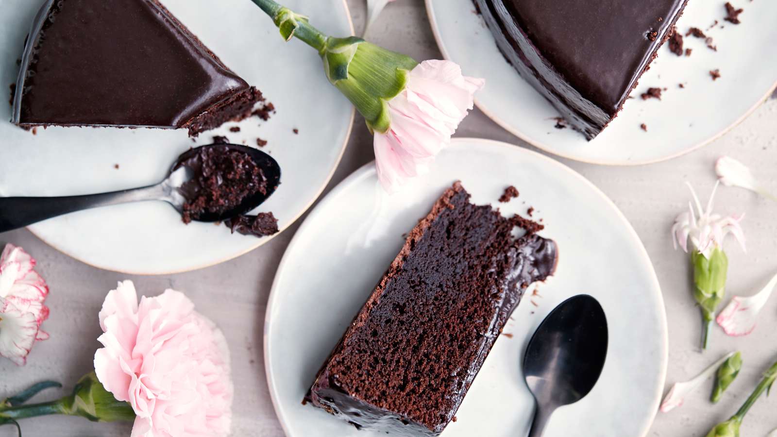 Kolme suklaakakkupalaa kakkulautasilla, kaksi lusikkaa ja vaaleanpunaisia neilikoita pöydän päällä.
