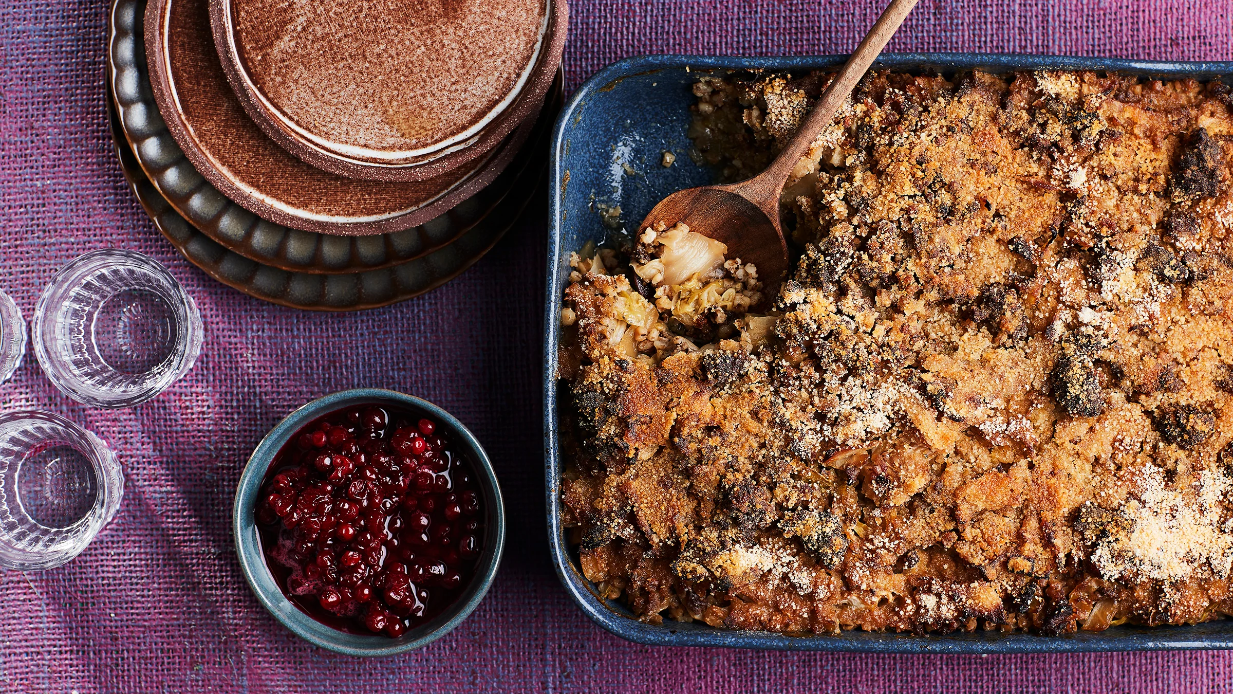 Uunivuoka, jossa kaalilaatikkoa ja puinen otin. Vuoan vierellä puolukkahilloa kulhossa. Kuvan reunalla osittain pois rajautuneena laseja sekä pino lautasia.