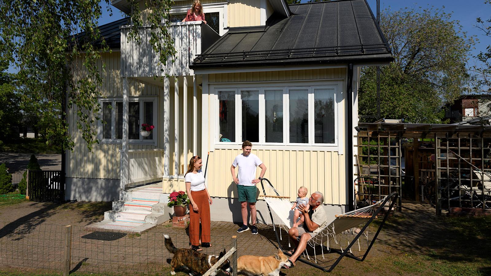 Perhe kuvattuna puutalon edessä kokoontuneena riippumaton viereen, kaksi koiraa vilistää pihalla