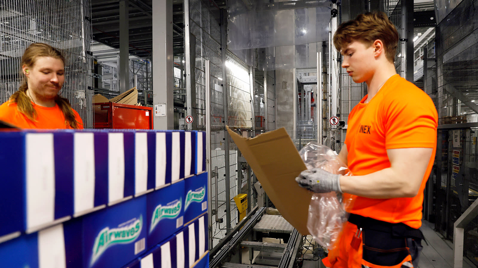 Kaksi työasuun pukeutunutta miestä logistiikkakeskuksessa. Toinen taittelee pahvia.
